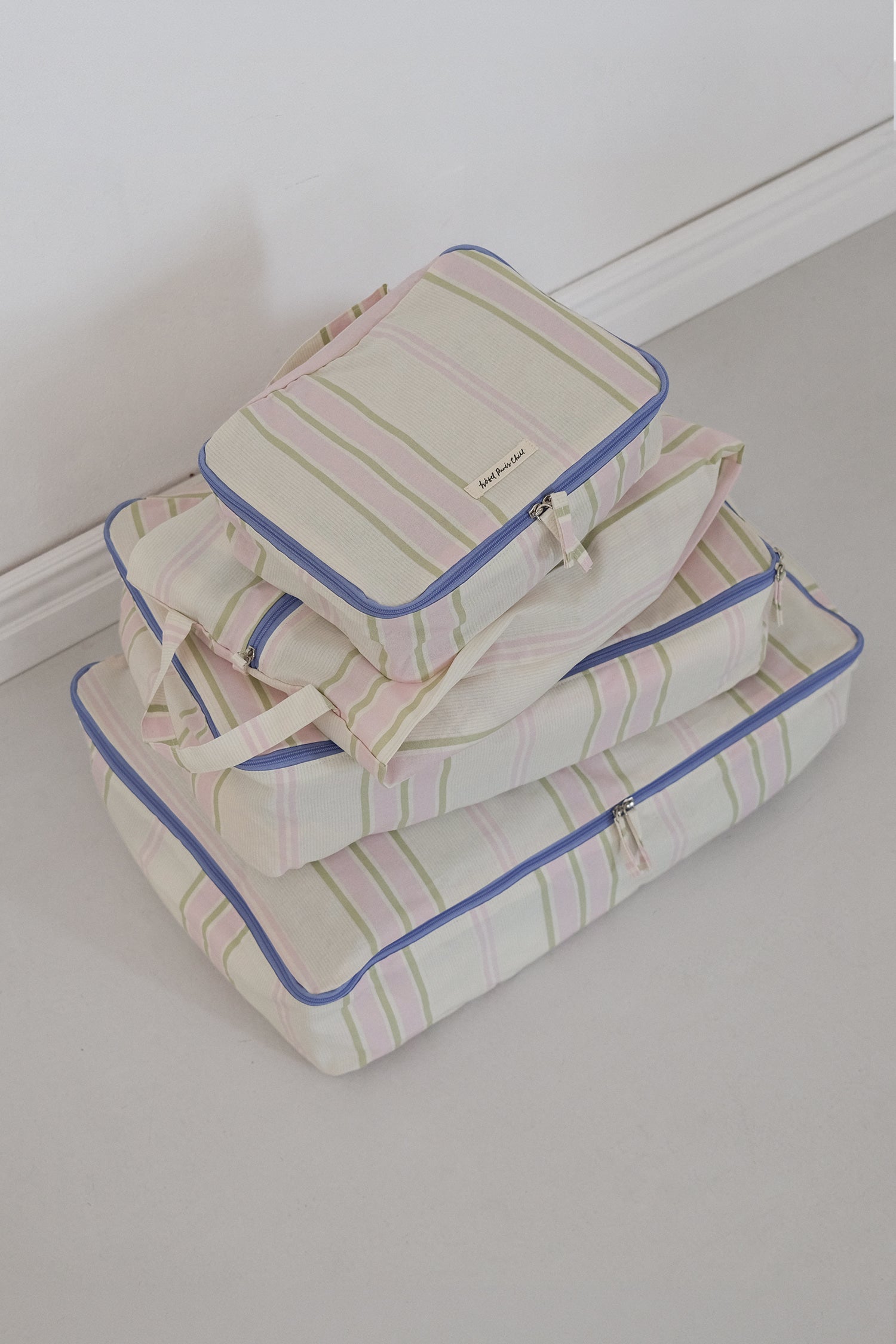 A stack of three striped Hotel827 cotton travel pouches in Lilac, shown on a light gray floor against a white wall.